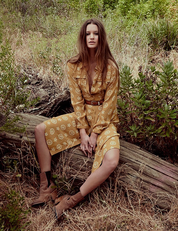 A woman in a patterned dress and boots sits on a log in a grassy field with scattered trees and foliage in an outdoor setting.