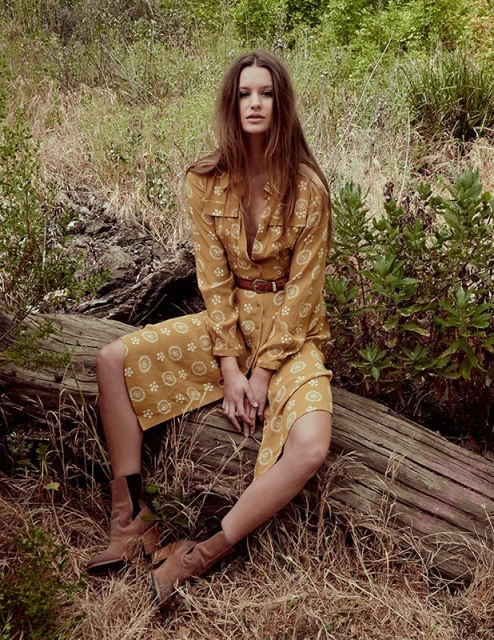 A woman in a patterned dress and boots sits on a log in a grassy field with scattered trees and foliage in an outdoor setting.