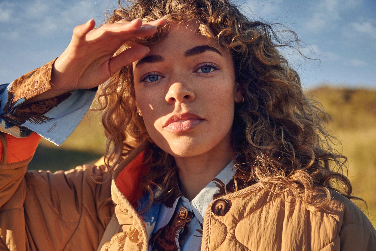 Close-up of an adult female with curly hair shielding her eyes with her hand, wearing a patterned blouse and tan quilted jacket.