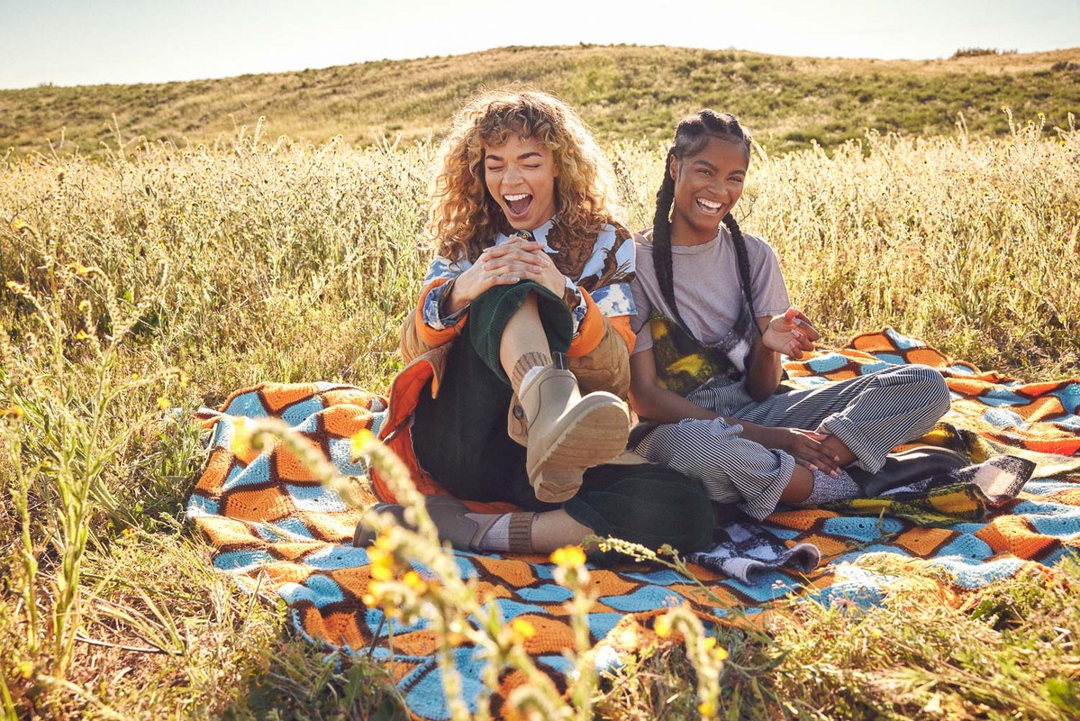 Two young women laughing and sitting on a colorful blanket in a grassy field on a sunny day, golden hour lighting, outdoors.