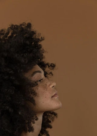 Profile portrait of a Black woman with voluminous curly dark hair and freckles against a neutral brown background