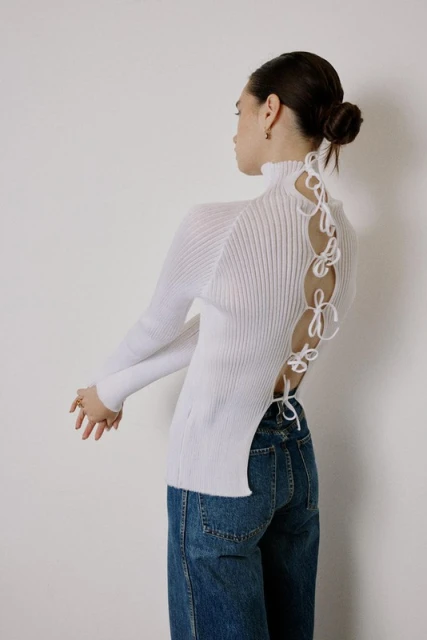 A young woman poses in a white ribbed turtleneck sweater with an open back tied with ribbons and wide-leg blue jeans against a plain background.