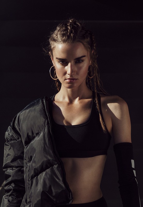 Portrait of a young adult woman in athletic wear with a puffer jacket slung over her shoulder against a dark background in studio lighting.