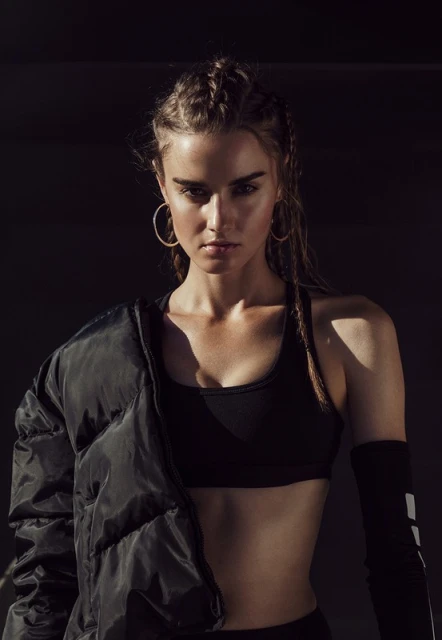 Portrait of a young adult woman in athletic wear with a puffer jacket slung over her shoulder against a dark background in studio lighting.
