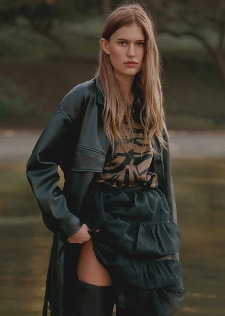 A fair-skinned woman with long blonde hair poses in a black leather jacket, a printed top, and a tiered black skirt in an outdoor setting.