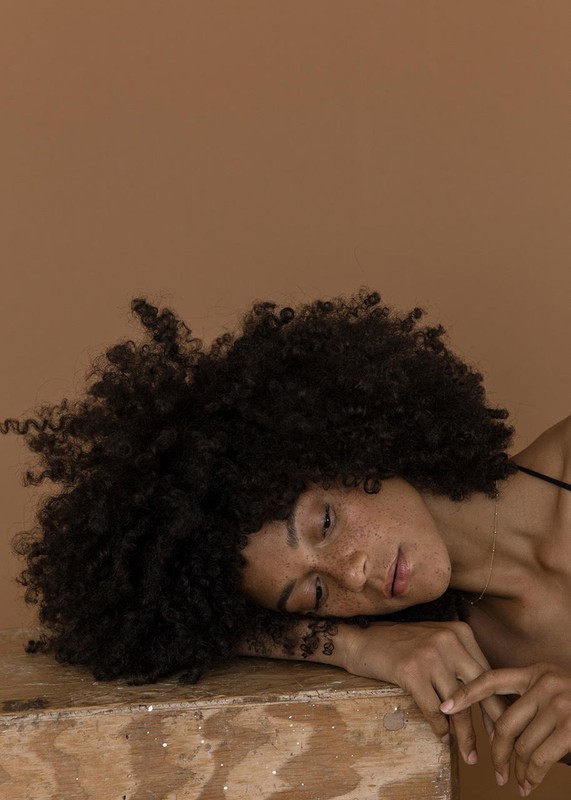 Portrait of a woman with afro resting her head on a wooden surface, set against a solid brown background in a photo studio setting.