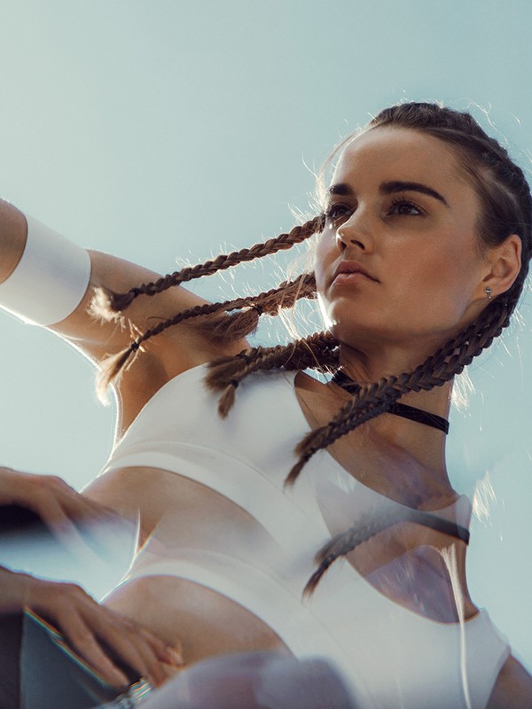 Low-angle shot of a woman with braided hair wearing a white sports bra and choker necklace, set against a clear sky with a blurry effect.