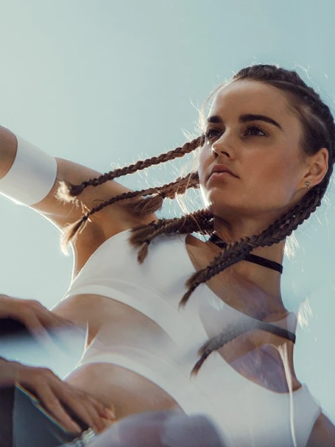 Low-angle shot of a woman with braided hair wearing a white sports bra and choker necklace, set against a clear sky with a blurry effect.
