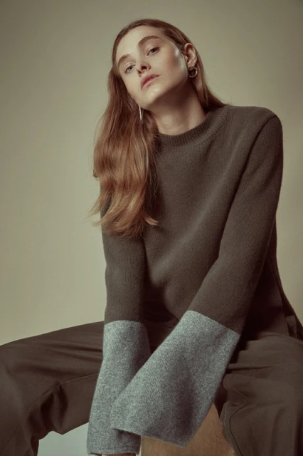 A young adult woman with long auburn hair poses indoors wearing a two-tone sweater and pants, seated against a neutral background.