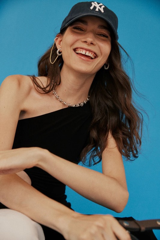 Smiling young woman with dark hair and a baseball cap wears a black one-shoulder dress against a solid blue studio backdrop.