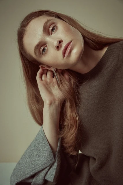 Portrait of a redhead woman in a minimalist fashion studio shot, wearing a brown sweater and silver earrings, looking pensively at the viewer.