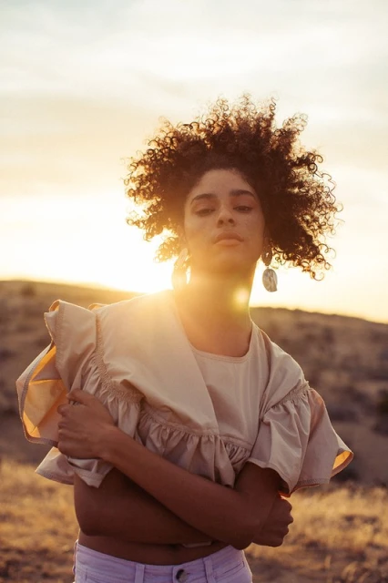 Portrait of a woman with curly hair wearing a ruffled top in a sunlit, natural setting, showcasing fashion and beauty.