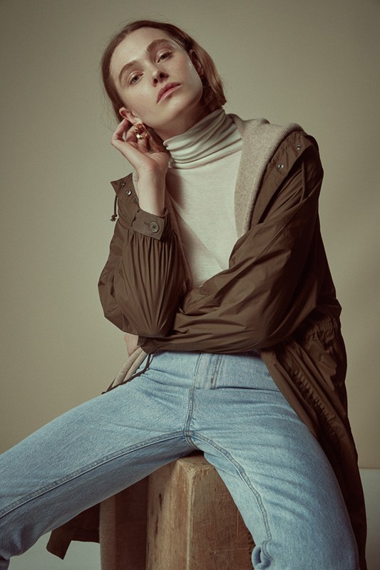 Portrait of a young fair-skinned woman with light brown hair, wearing a tan turtleneck, blue jeans, and brown coat sitting on a wooden box.