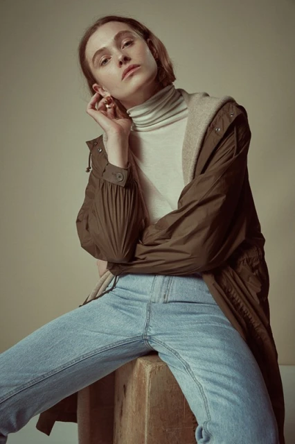 Portrait of a young fair-skinned woman with light brown hair, wearing a tan turtleneck, blue jeans, and brown coat sitting on a wooden box.