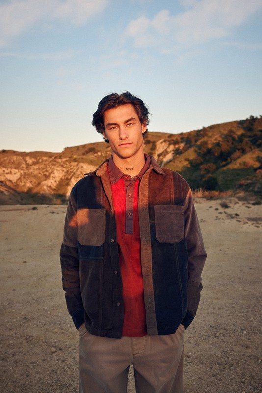 A portrait of a young adult male outdoors, wearing a multi-panel jacket and red collared shirt, posing against a background of hills and sky.