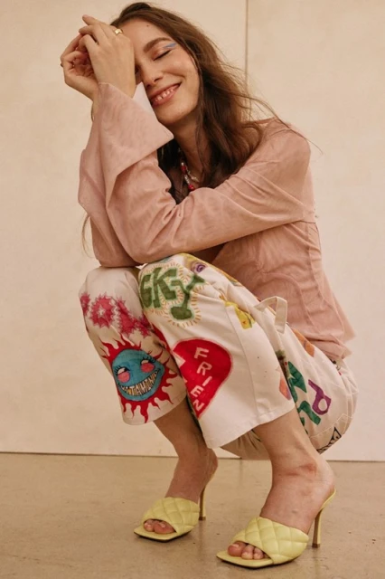 A fair-skinned young woman with long brown hair sits smiling in a pink top and patterned pants, wearing yellow high-heeled shoes, indoors.