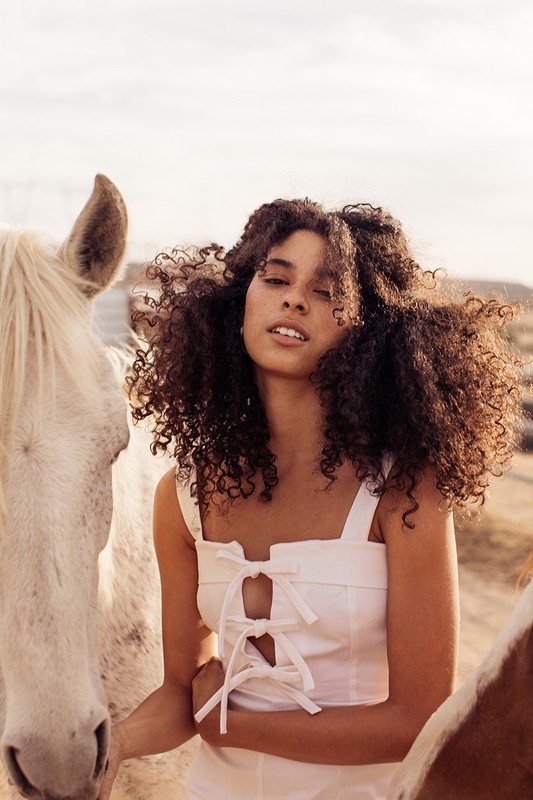 Young woman with curly hair poses with two horses outdoors on a sunny day wearing a white tie-front top, creating a natural, serene scene.
