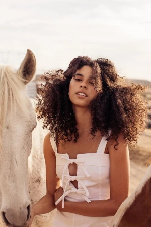 Young woman with curly hair poses with two horses outdoors on a sunny day wearing a white tie-front top, creating a natural, serene scene.