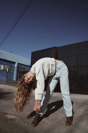 A woman with long wavy blonde hair bending backwards outdoors in Los Angeles, wearing a cream sweater and light blue jeans