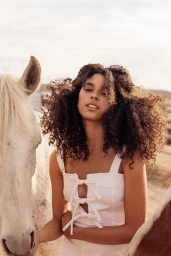 A woman with voluminous curly brown hair wearing a white tie-front top stands outdoors between two horses.