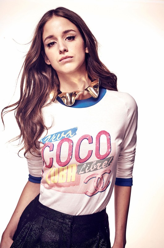 Portrait of a young adult woman in a long-sleeved graphic tee and shorts, accessorized with a statement necklace, set against a bright backdrop.