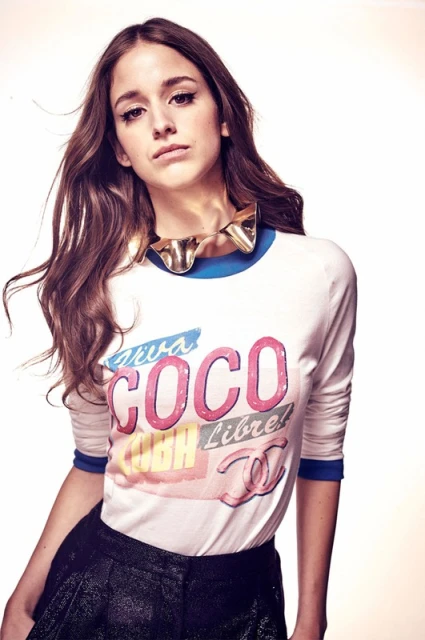 Portrait of a young adult woman in a long-sleeved graphic tee and shorts, accessorized with a statement necklace, set against a bright backdrop.