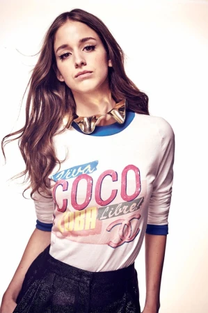 Portrait of a young adult woman in a long-sleeved graphic tee and shorts, accessorized with a statement necklace, set against a bright backdrop.
