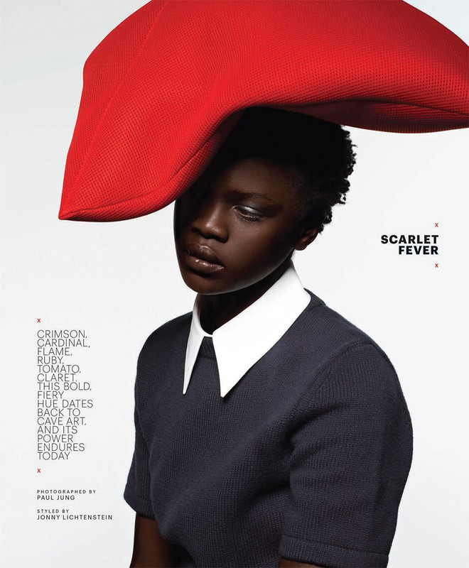 Fashion portrait of a black woman with short hair wearing a dark top with white collar and large red hat, against a bright background.