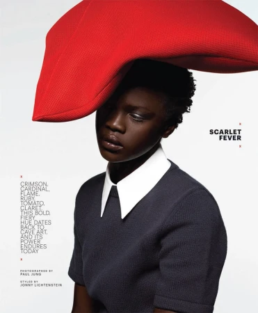 Fashion portrait of a black woman with short hair wearing a dark top with white collar and large red hat, against a bright background.