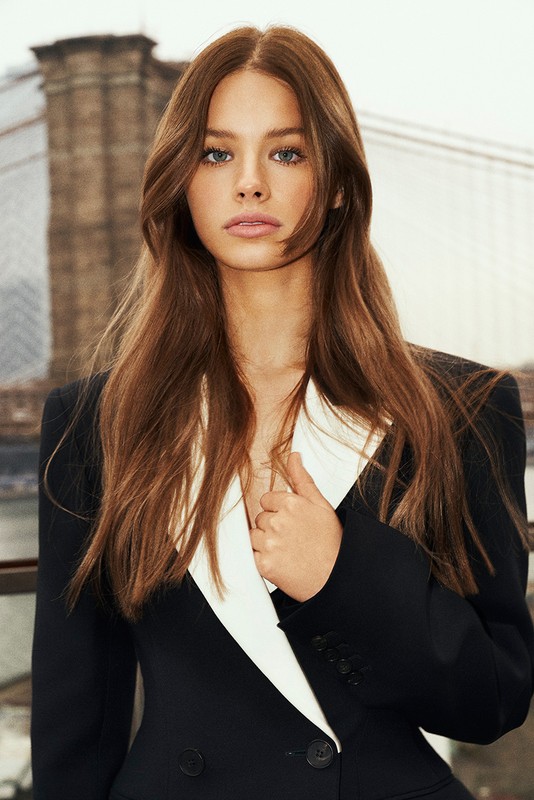 Close-up of a woman with long brown hair wearing a black and white blazer in front of an out-of-focus urban background.