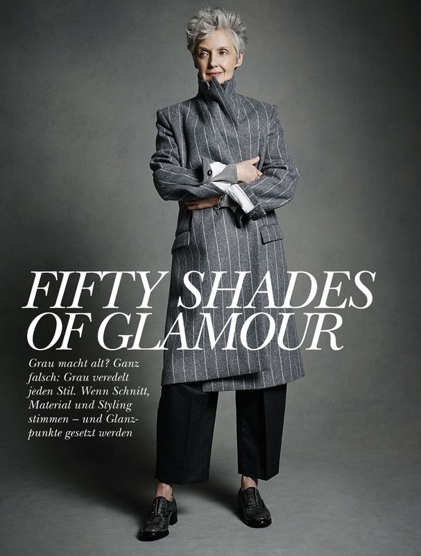 An elderly woman stands in a gray coat and black pants with arms crossed in a fashion editorial image with gray background.