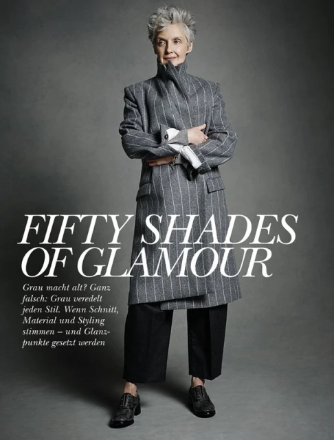 An elderly woman stands in a gray coat and black pants with arms crossed in a fashion editorial image with gray background.