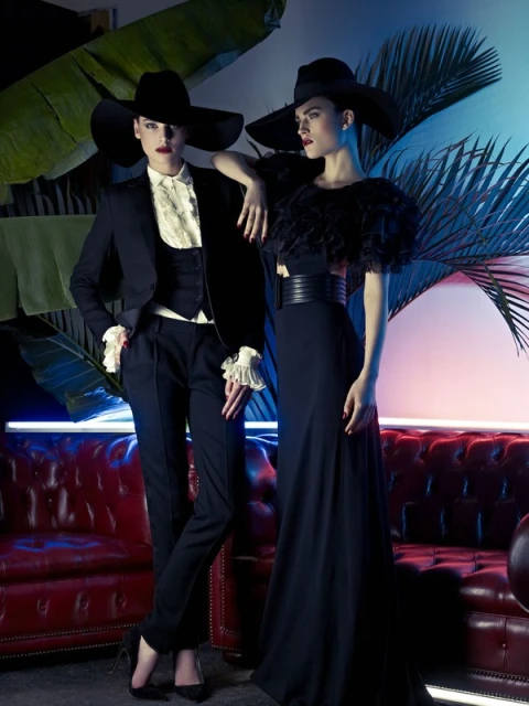 Two women pose in elegant black dresses and hats against a backdrop of tropical foliage and ambient blue and pink lighting. 