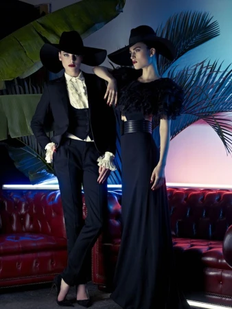 Two women pose in elegant black dresses and hats against a backdrop of tropical foliage and ambient blue and pink lighting. 