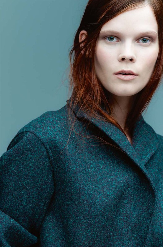 Close-up fashion portrait of an adult female with red hair wearing a green wool coat against a pale blue background in a studio setting.