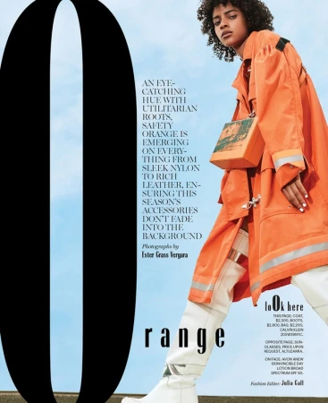 Fashion editorial shot of a woman wearing an orange coat and white boots, posing in front of a blue sky with a large black letter.