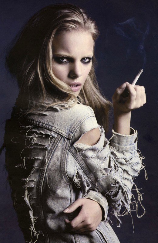 Blonde woman in distressed denim jacket smokes a cigarette against a dark background, gazing intensely at the camera.