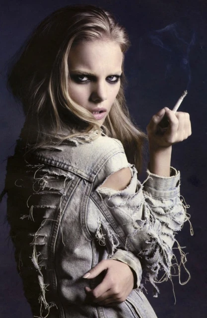 Blonde woman in distressed denim jacket smokes a cigarette against a dark background, gazing intensely at the camera.