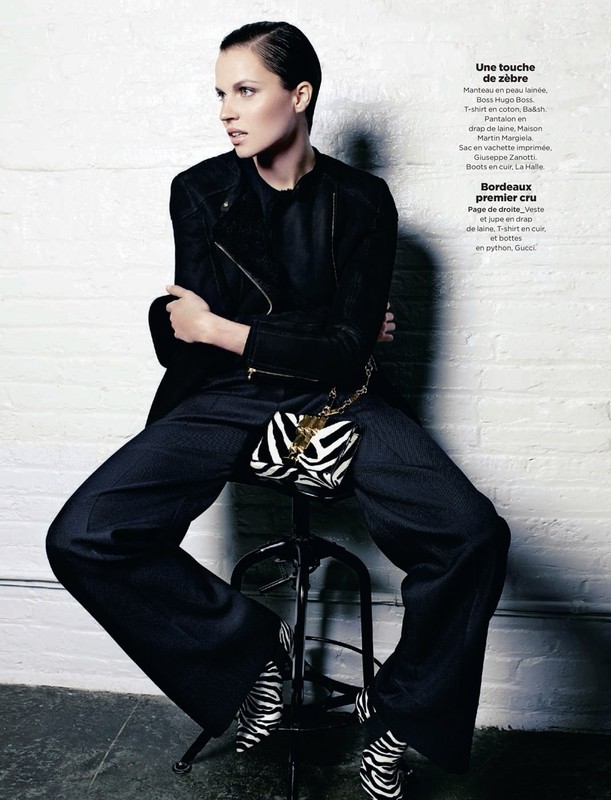 Fashion editorial shot of a woman in a black jacket and wide-leg pants with a zebra-patterned purse and heels, seated indoors.