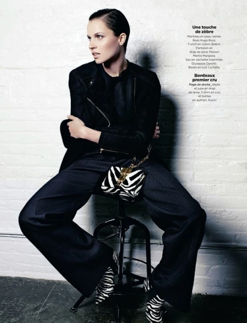 Fashion editorial shot of a woman in a black jacket and wide-leg pants with a zebra-patterned purse and heels, seated indoors.