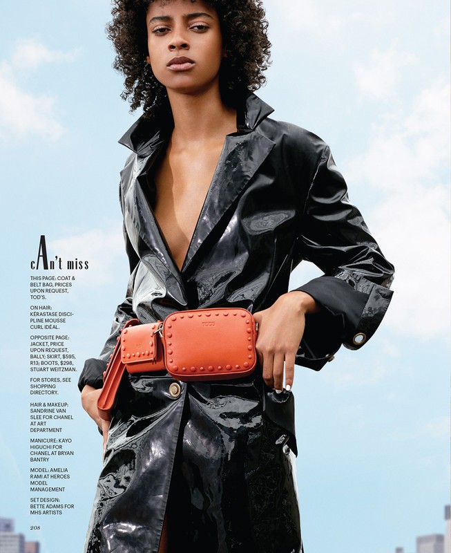 Fashion editorial shot of a woman in a black trench coat with an orange belt bag, posing against a light blue sky and a city skyline.
