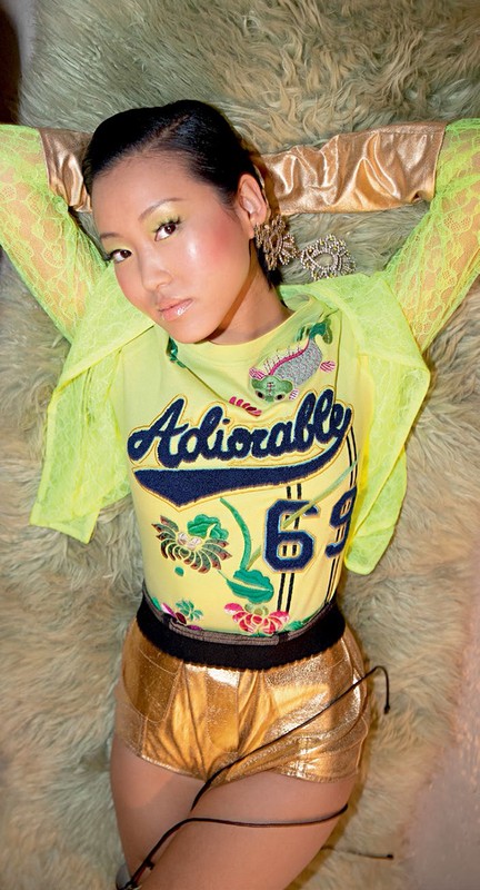 A woman poses indoors wearing a yellow top, gold shorts, and statement earrings, lying on a furry, light-green surface with hands behind head.