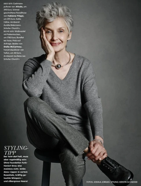 Elegant woman with gray hair sits on a stool, posing in a gray sweater and pants. She wears a silver necklace and bracelet.