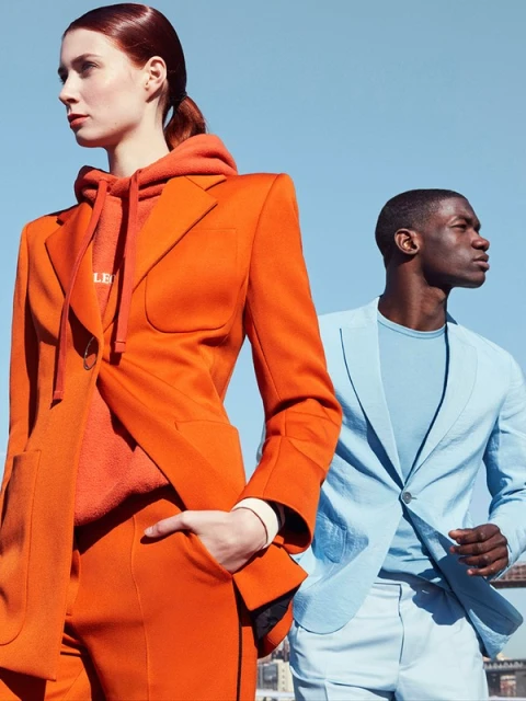 Two fashion models wearing blazers, posing outdoors against a bright blue sky; one in orange and one in light blue.