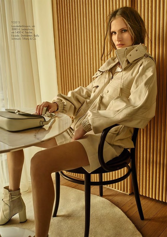Fashion shot of a woman in a beige jacket and skirt sitting at a table with a purse, set against a wooden slat wall.