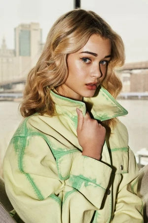 Close-up portrait of a blonde woman wearing a light yellow jacket with green contrast stitching, with a city skyline visible in the background.