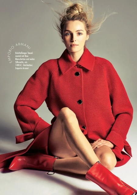 Blonde fashion model in red overcoat and matching boots sits in a studio pose. Fashion editorial for outerwear. Studio shot.