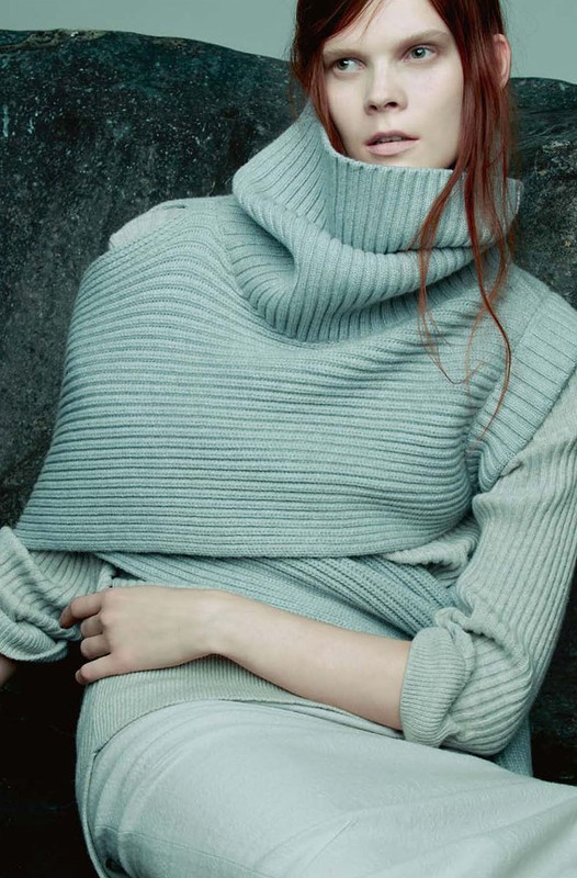 A fair-skinned young adult woman with red hair poses in a light blue sweater and skirt, next to a rock in a studio setting.
