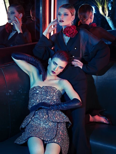 Two adult female fashion models pose in a dimly lit indoor environment, wearing evening wear, long gloves, and rose chokers.