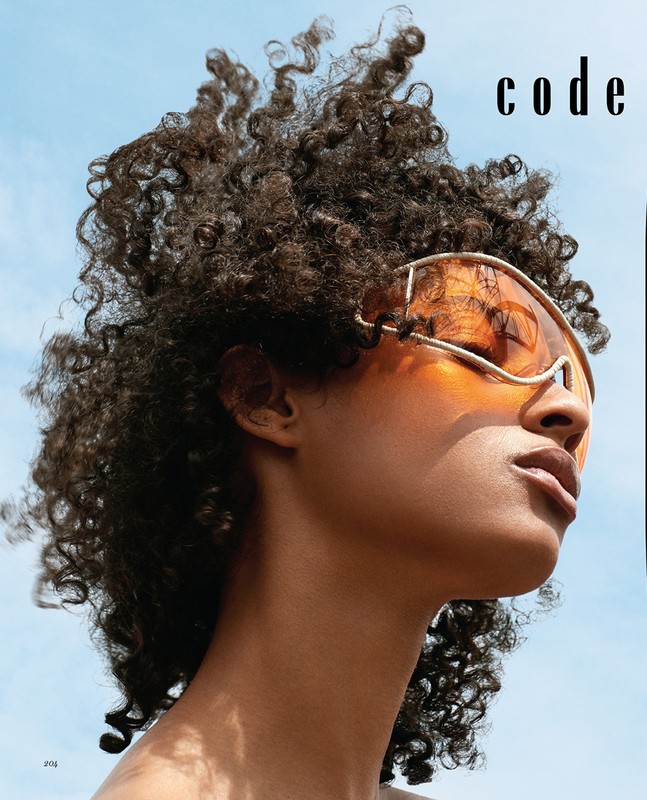 Close-up of a Black female model with an afro wearing futuristic orange glasses against a light blue sky, fashion magazine cover.